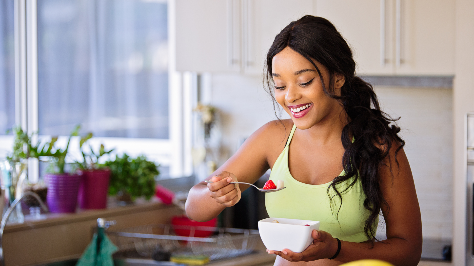 A woman in eating a spoonful of fruits against a kitchen background.