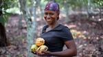 A smiling woman standing in a cocoa plantation, holding ripe cocoa pods in her hands. She is wearing a patterned headscarf and a black shirt.