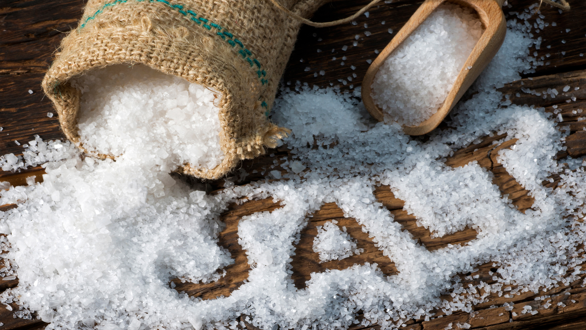 Coarse salt spilled from burlap sack with salt written on wooden surface.