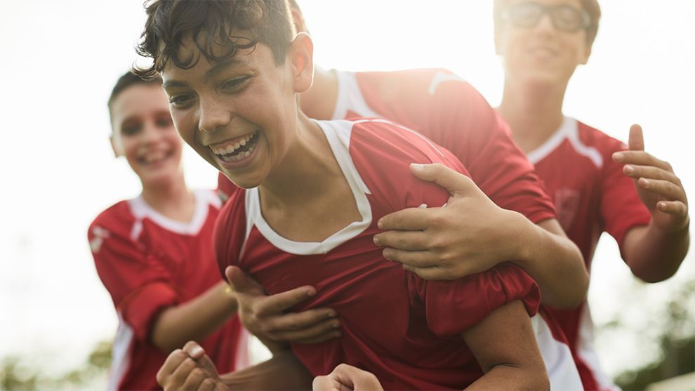 Four young boys in matching red shirts, smiling and hugging after scoring a goal.