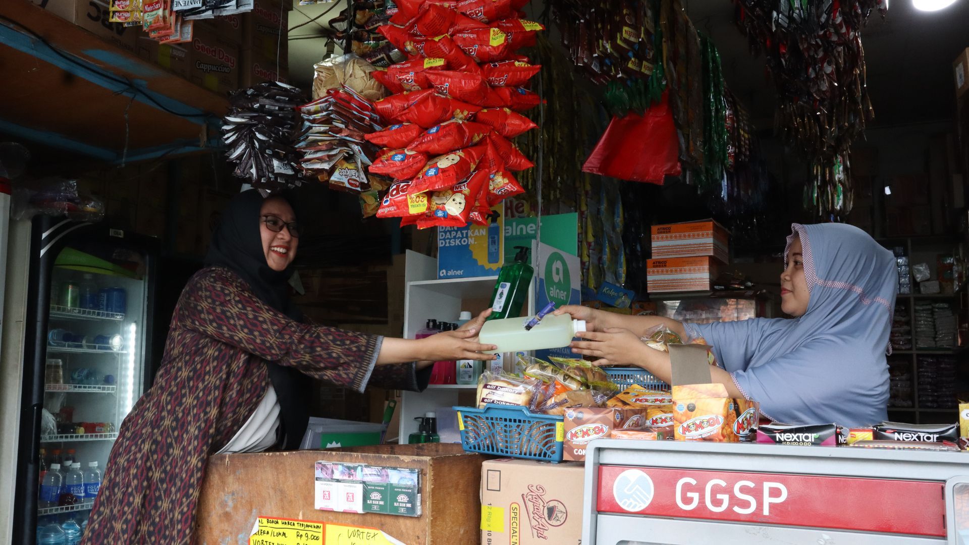 Two women in hijabs smiling at each other in a shop whilst one passes the other a bottle of product.