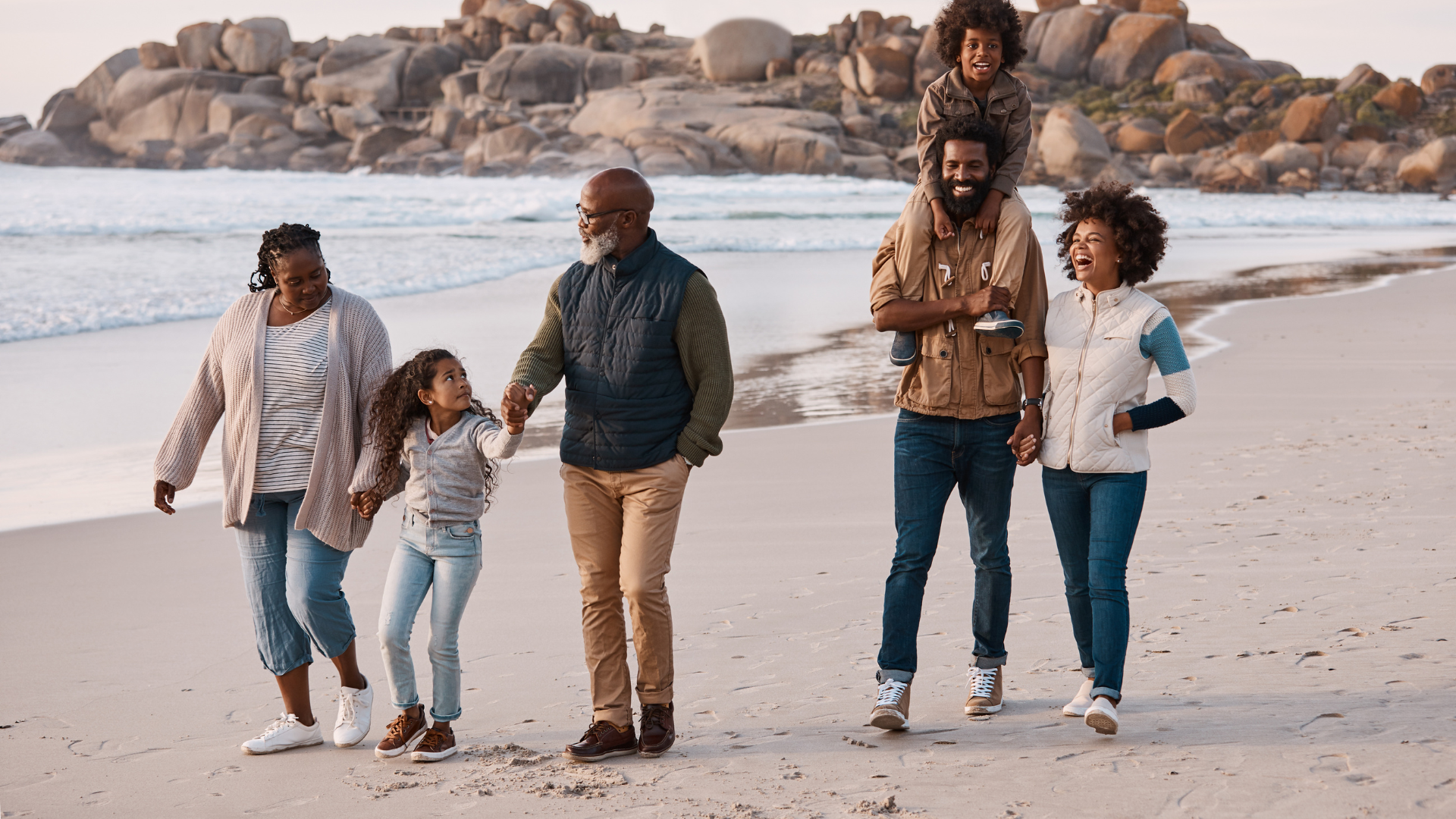 Family walking on a sandy beach with rocky background.