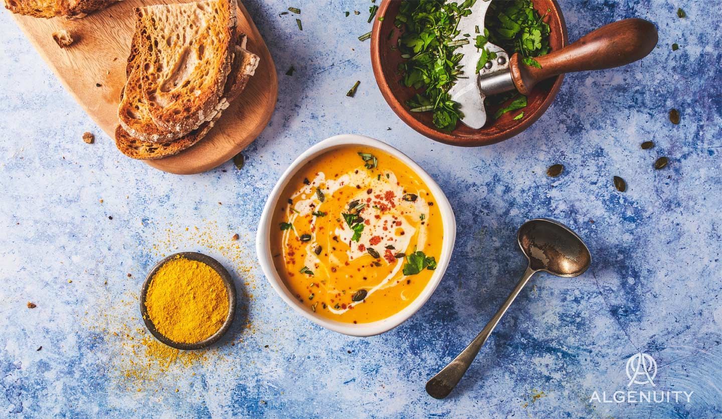 A bowl of orange soup, a small bowl of orange microalgae powder, a bowl of green herbs and a board with sliced brown bread.