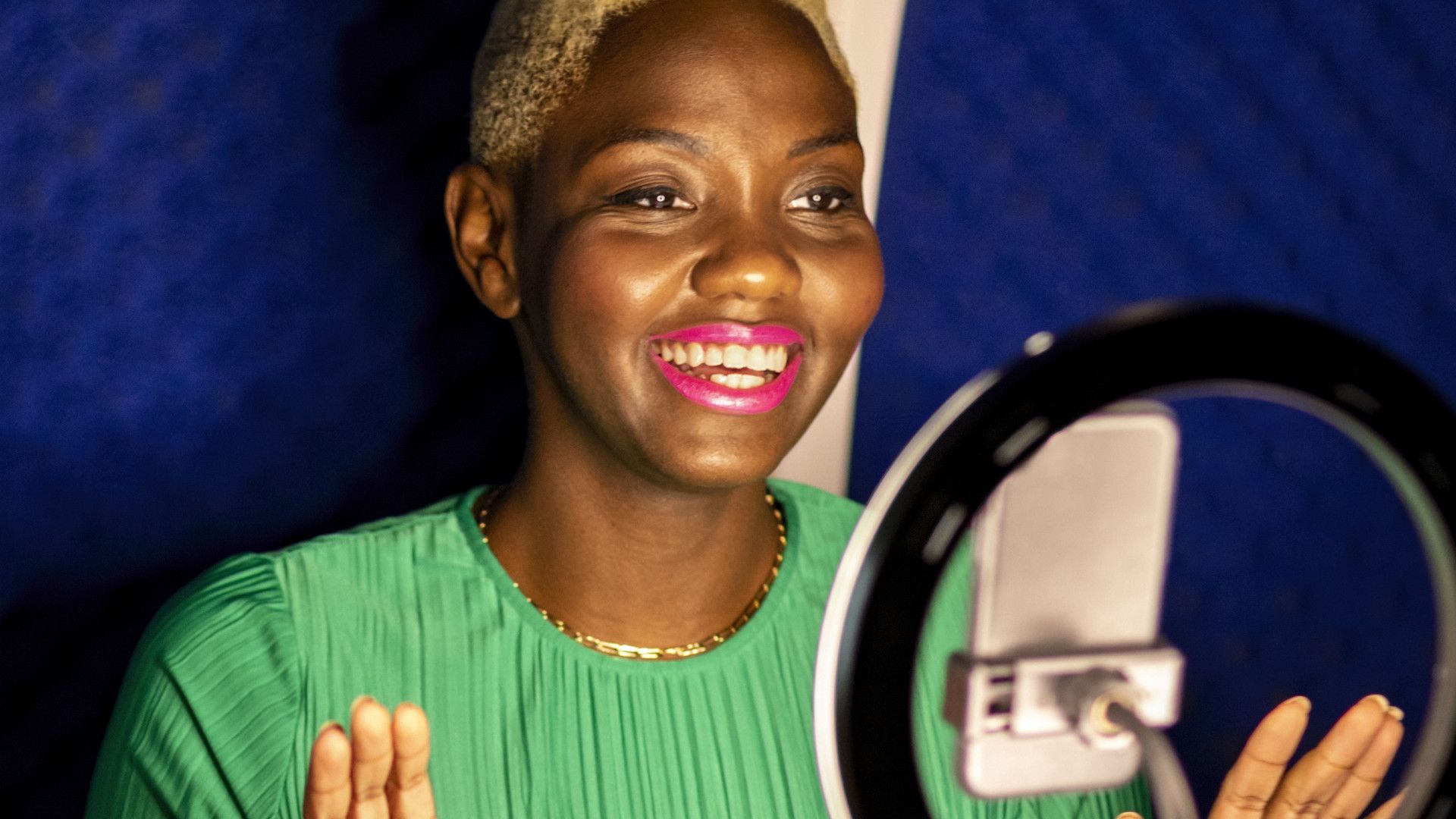 A young Black woman with bleached, short hair and a vivid green dress smiles at a camera. A ring light is also in the frame.