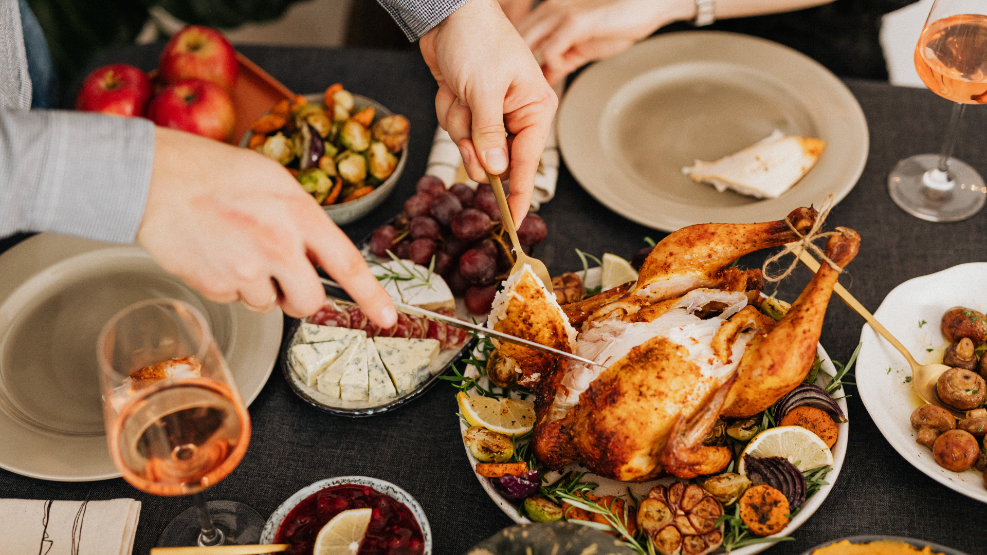 Person carving a roasted chicken on a table with sides and wine.