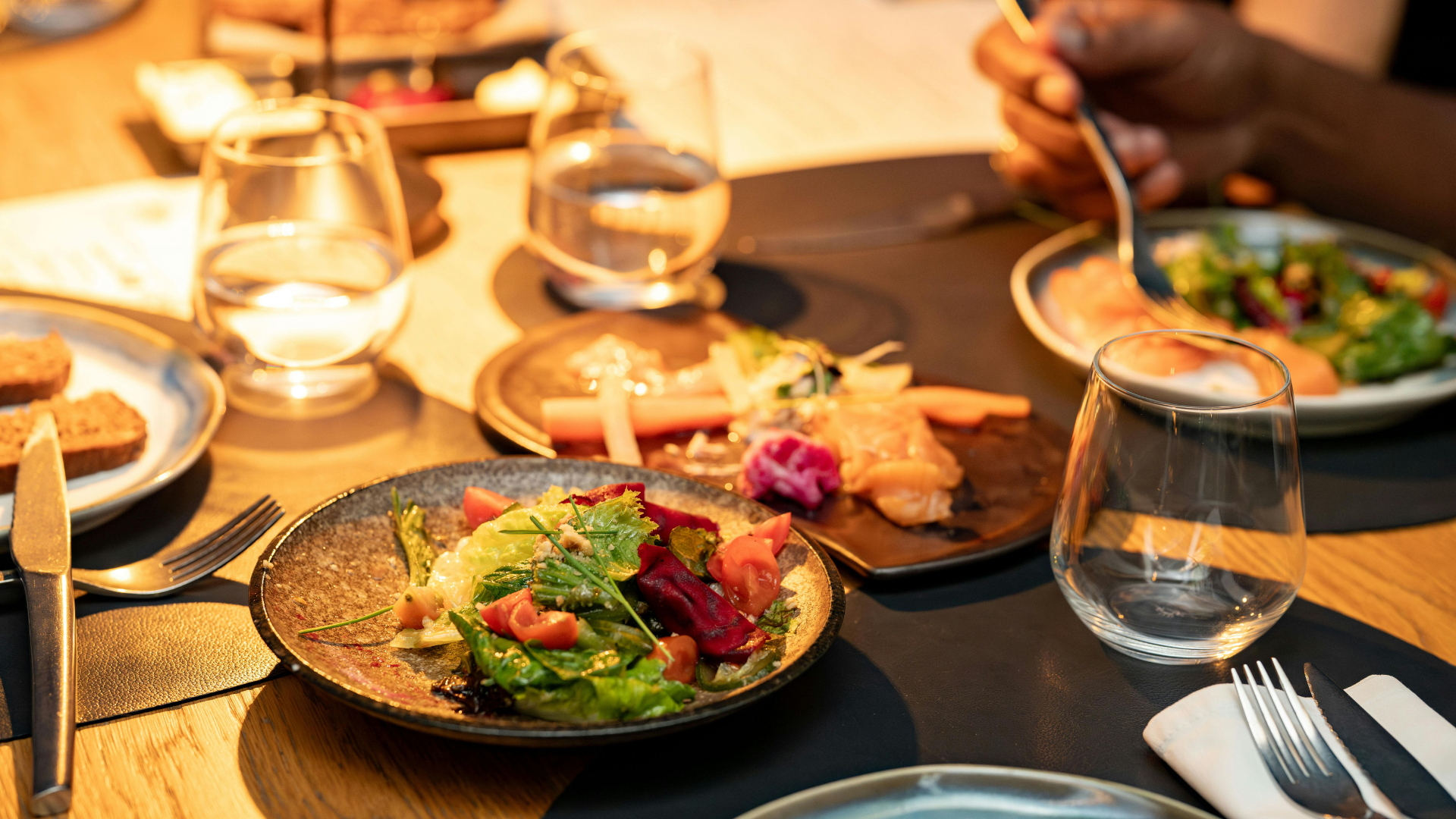 A dinner table featuring colorful salads, bread, and water glasses and utensils, set in a warm, inviting atmosphere.