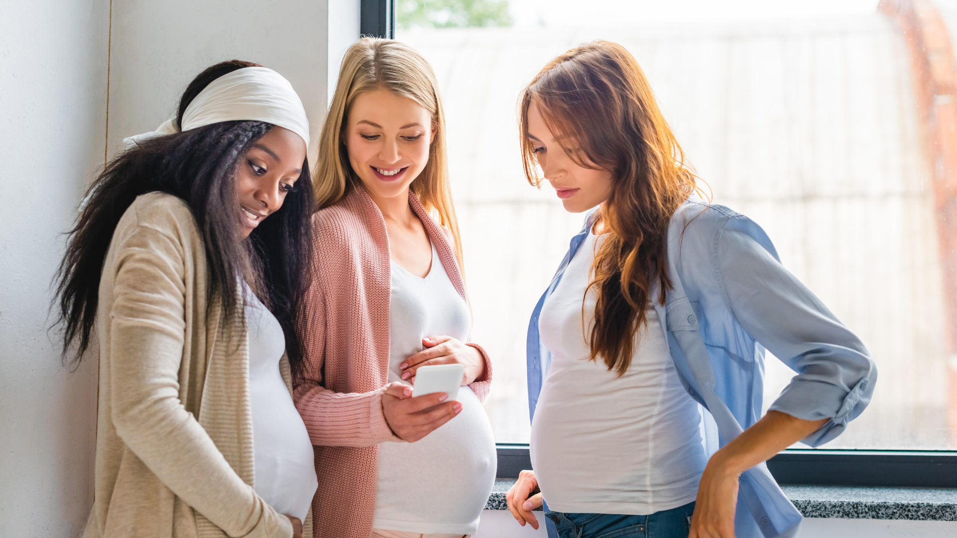Three pregnant women stand together, smiling and looking at a smartphone.