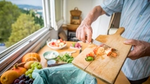 A person slices vegetables on a wooden cutting board, with a green trash bag nearby for food scraps and various fruits on the table.