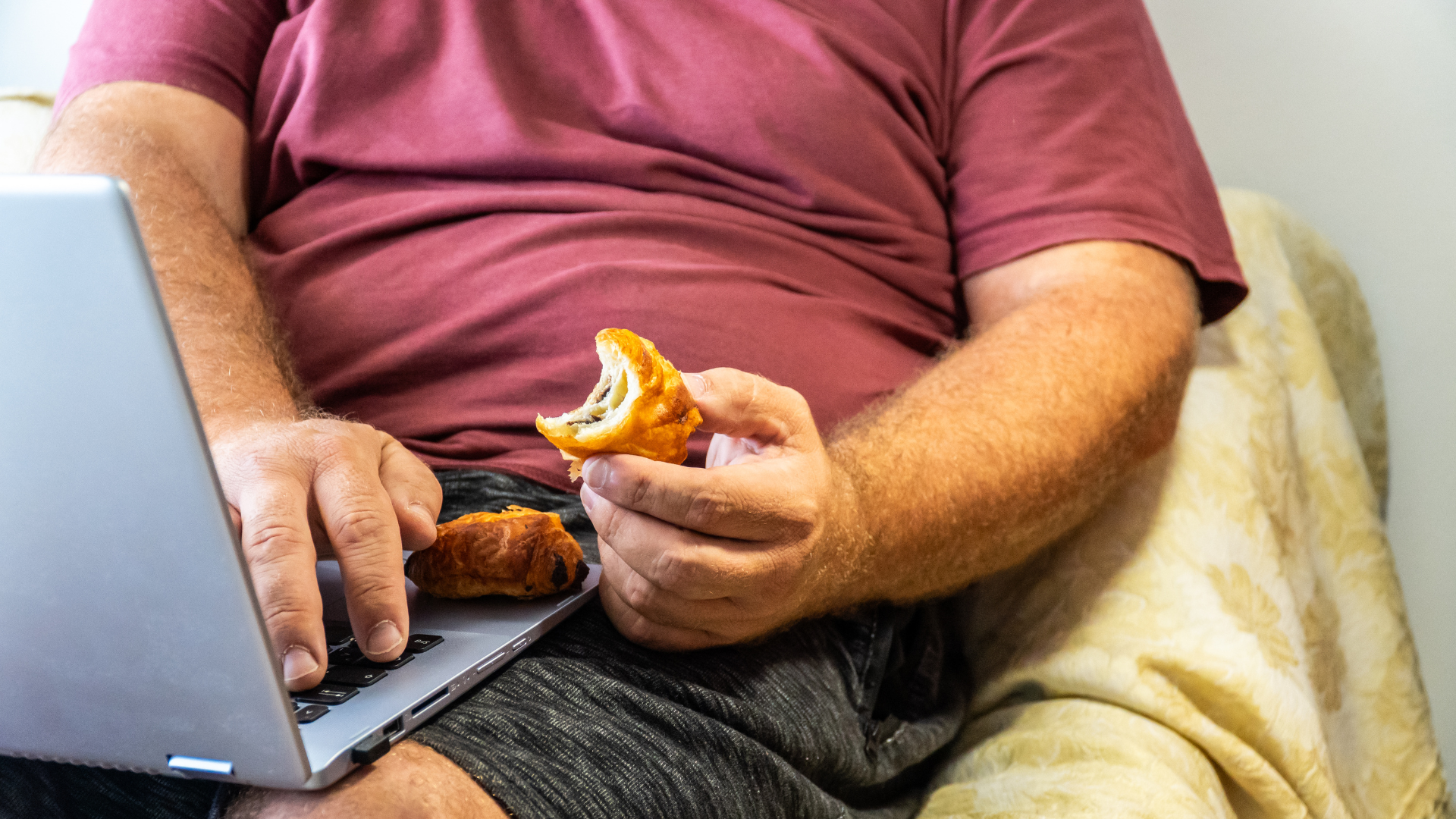 Person eating pastry while using a laptop on a couch.