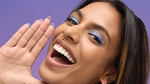 A woman with long dark hair and blue eye makeup smiles brightly against a purple background. She holds one hand up beside her face, showcasing her white teeth and happy expression.