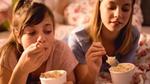 Two teenage girls lie on a bed eating big tubs of ice cream